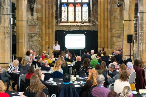 Groups of people sitting around several table looking towards a speaker in the distance. This is all in a converted church. 