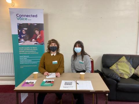 Jade and Carrie sit at a table in front of a pop Haref banner at a local community event