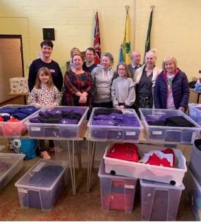 A group of 10 people of all ages standing in a church hall behind plastic boxes filled with clothes