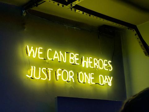 A wall-mounted neon sign spelling out the lyrics "WE CAN BE HEROES JUST FOR ONE DAY"